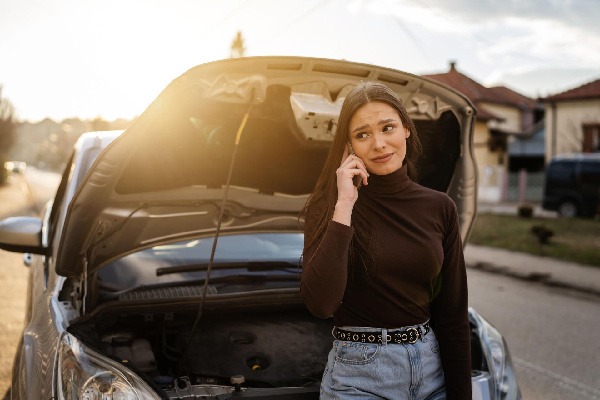 One young adult caucasian woman standing by her vehicle with open hood broken failed engine holding a phone calling towing service for help on the road Roadside assistance concept autumn or spring day